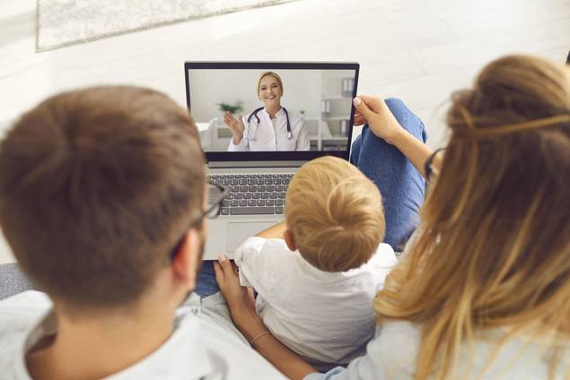 Ein Ehepaar sitzt mit seinem kleinen Kind auf einer Couch und führt ein Videotelefonat mit einer Ärztin, die lächelt und über einen Laptop-Bildschirm gestikuliert.