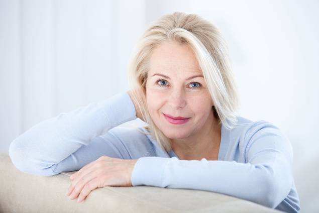 Eine lächelnde Frau mittleren Alters mit blondem Haar und blauen Augen, die einen hellblauen Pullover trägt, stützt ihren Arm auf die Rückenlehne einer beigen Couch in einem hellen, schwach beleuchteten Raum.