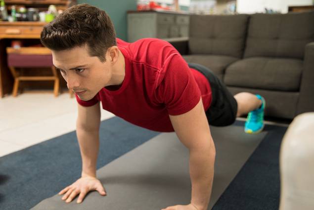 Ein Mann in einem roten Hemd und blauen Turnschuhen macht Liegestütze auf einer Yogamatte in einem Wohnzimmer, konzentriert und nach vorne blickend. Im Hintergrund sind eine Couch und ein Schreibtisch zu sehen.
